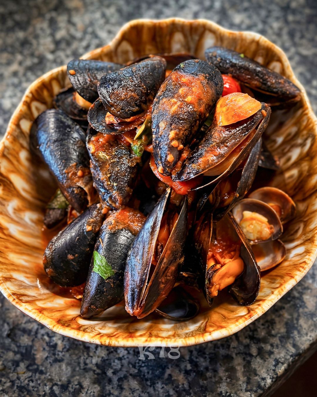 Una ciotola di cozze cotte con salsa piccante su un piano di lavoro maculato.