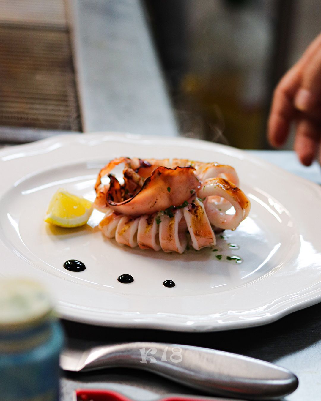 Frutti di mare grigliati serviti con spicchio di limone e salsa su un piatto bianco, in un ambiente da ristorante.