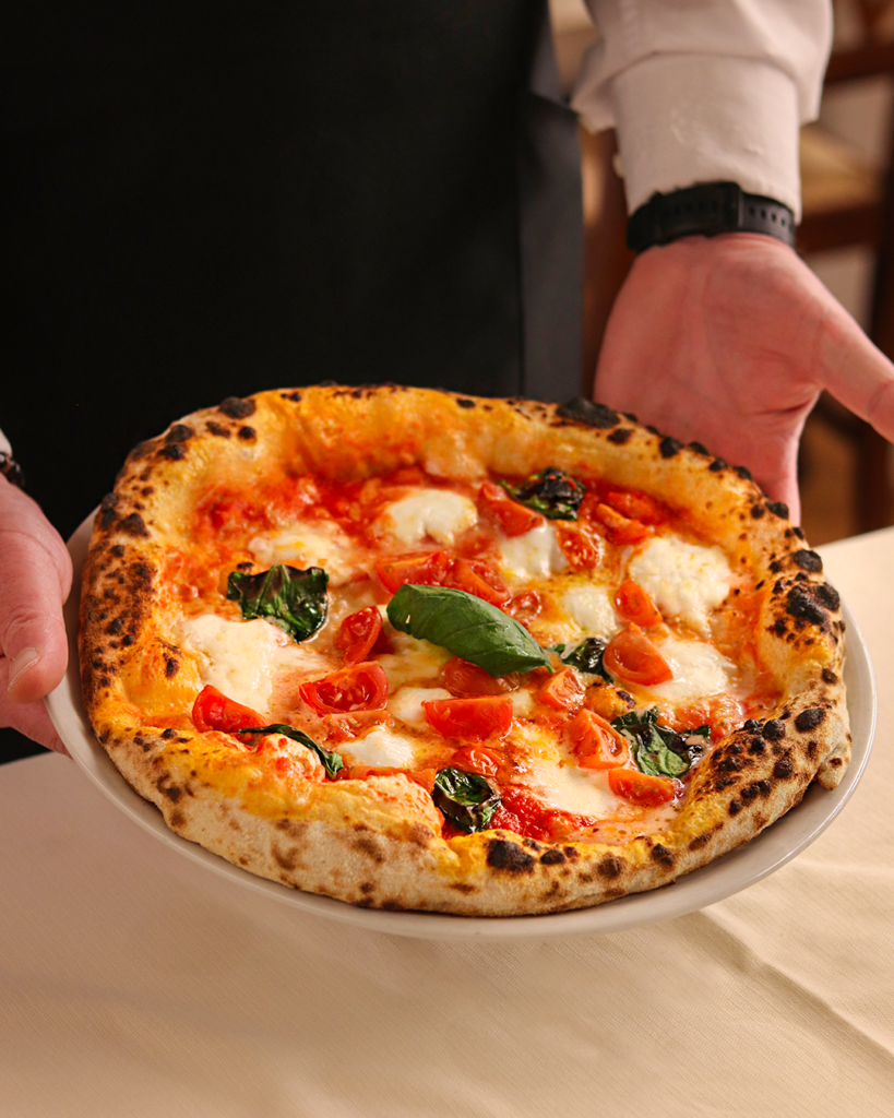 Mani che reggono una pizza margherita su un piatto, con formaggio fuso, salsa di pomodoro e foglie di basilico.