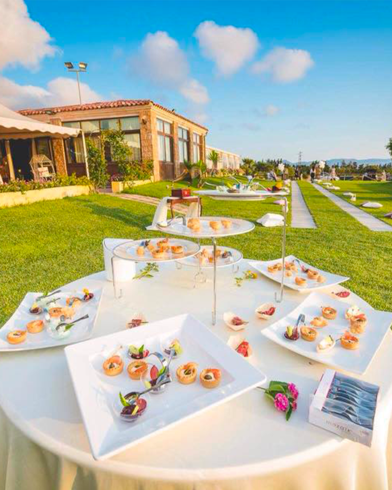 Buffet di dolci all'aperto su tavoli bianchi, accanto a un prato e a un edificio, sotto un cielo soleggiato.