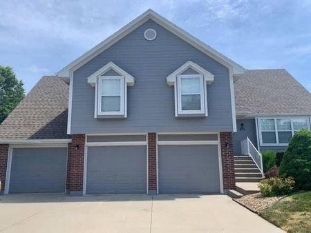 Two-story house with gray siding, three-car garage, and brown roof under a partly cloudy sky.