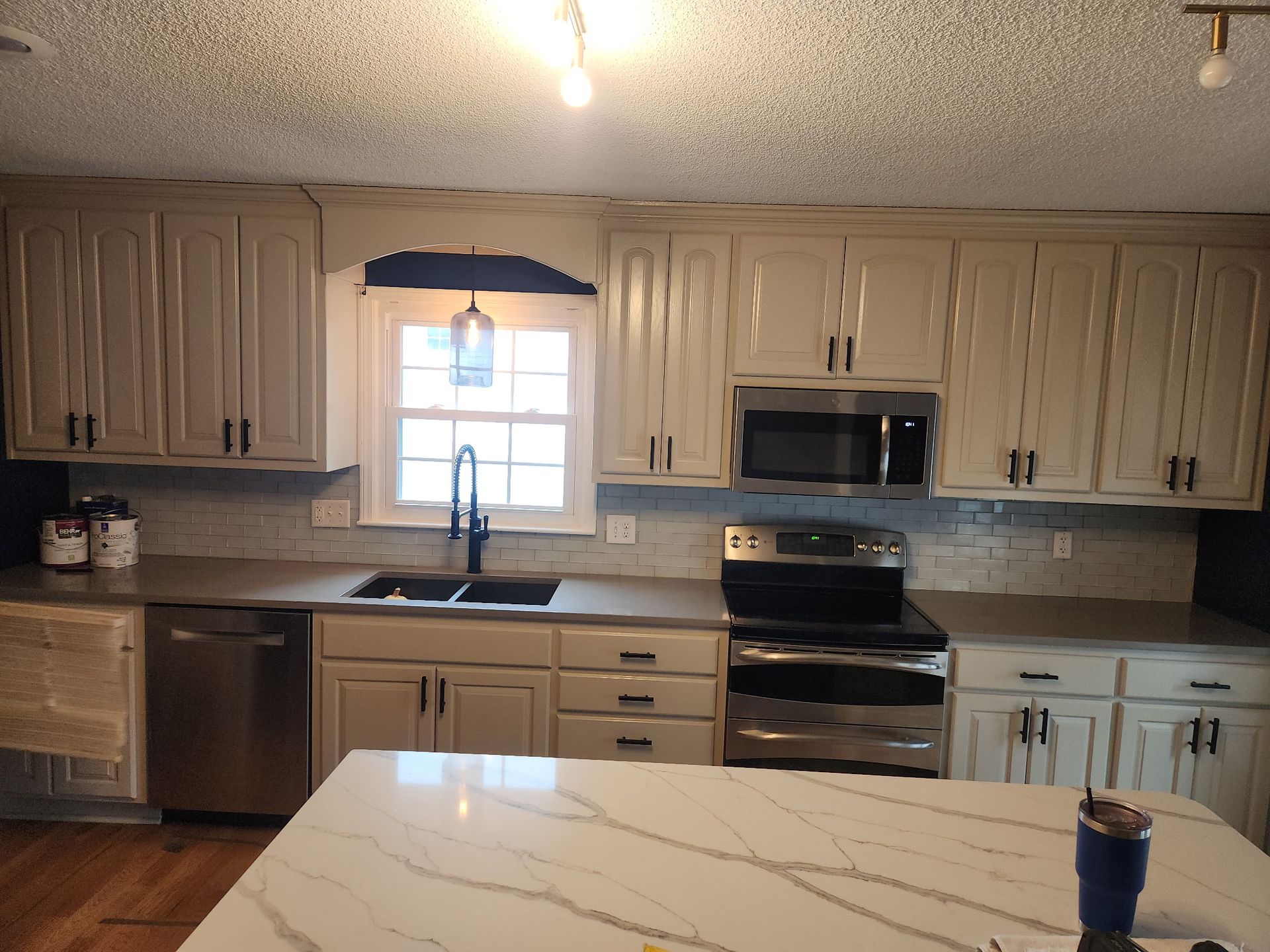 Kitchen with cream-colored cabinets, stainless steel appliances, a white marble-patterned island, and a sink by a window.