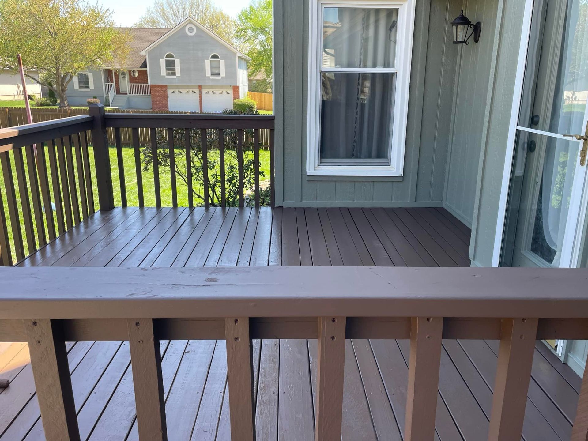 Brown wooden deck with railings, green siding, and a view of a house and yard.