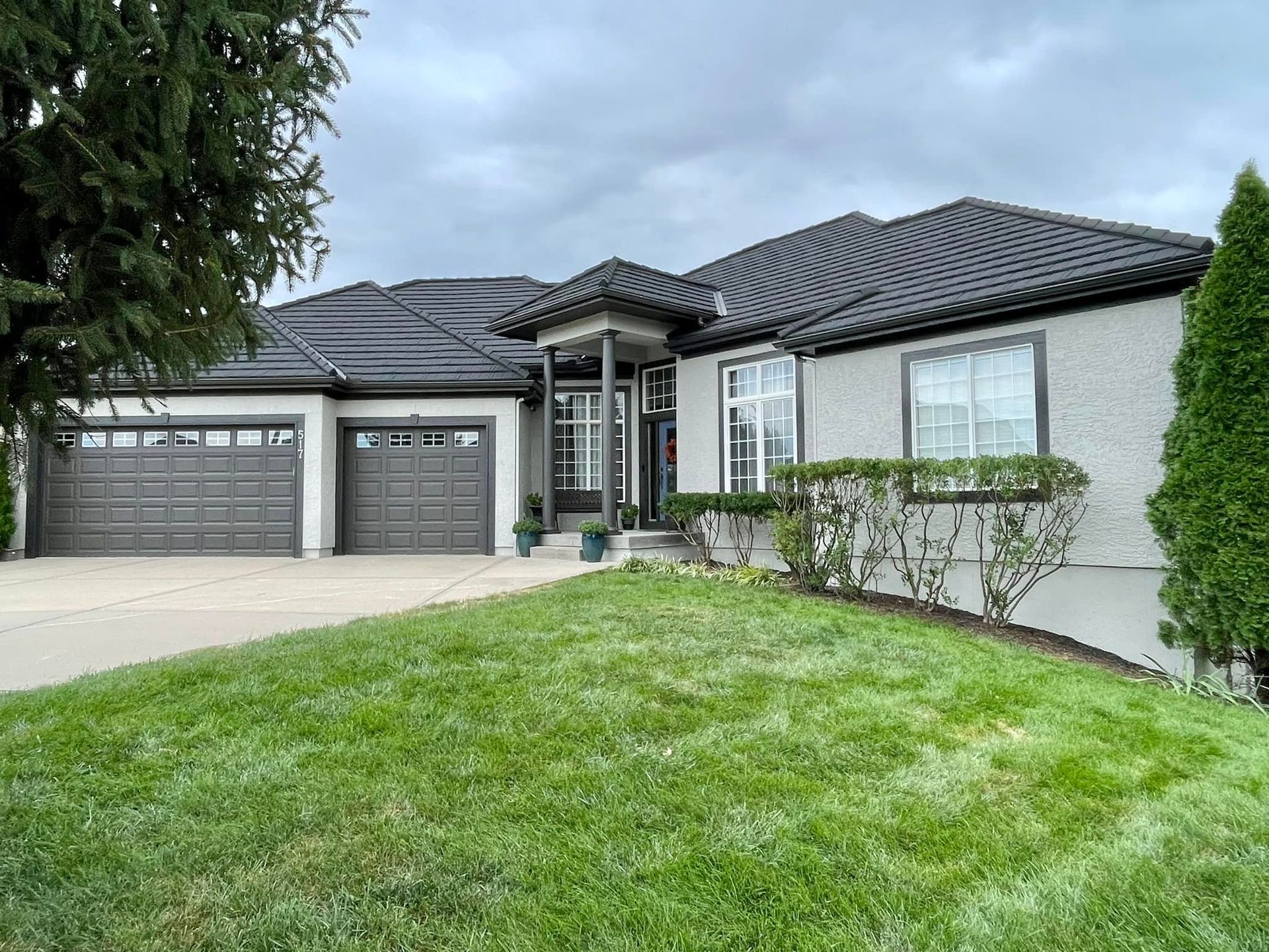 Gray stucco home with dark gray roof and garage doors, green lawn, cloudy sky.