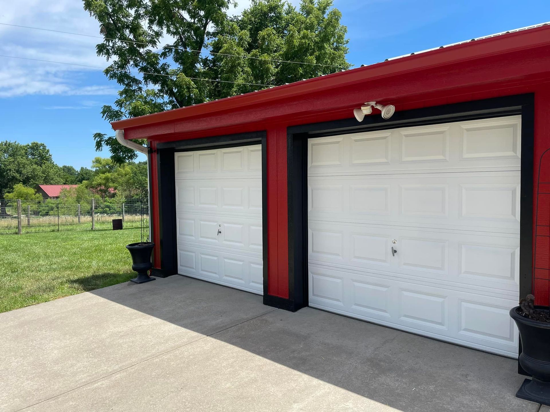 Two-car garage with white doors, red trim, black outlines, and a concrete driveway. Trees in the background.