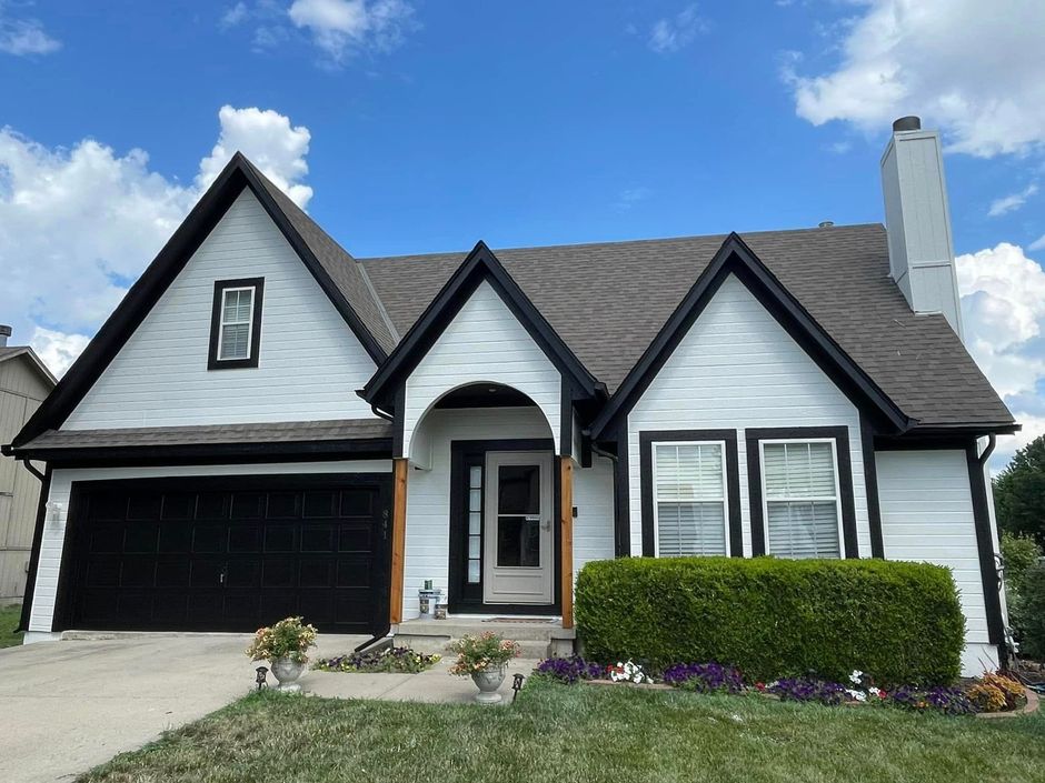 White house with black trim, garage door, and roof. Green shrubs and flowerbeds in front.