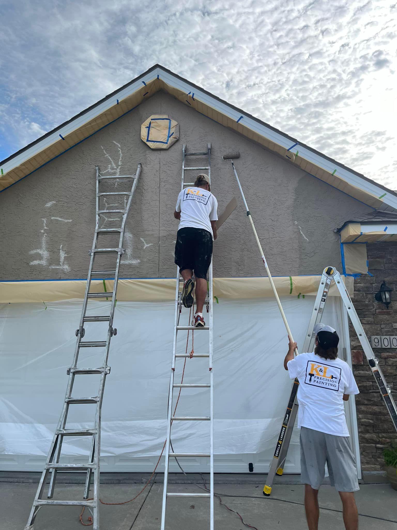 Three people painting a house exterior, with ladders against a tan gable wall.
