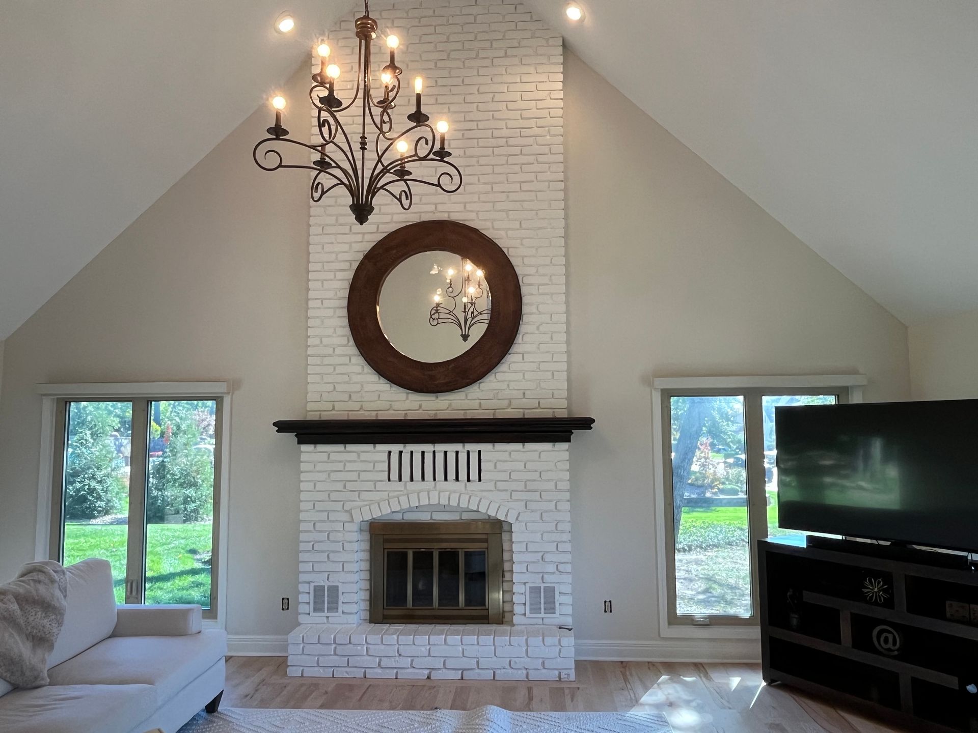 A living room with a white-painted brick fireplace, a dark wood-framed mirror, a chandelier, and a television on a stand.