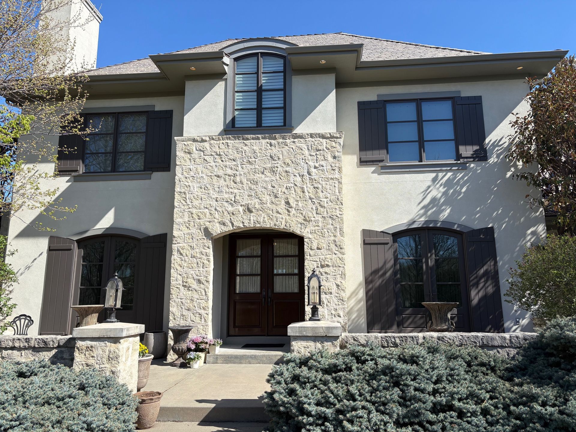 Two-story stucco house with stone facade, dark-framed windows, and a front entry surrounded by shrubs.
