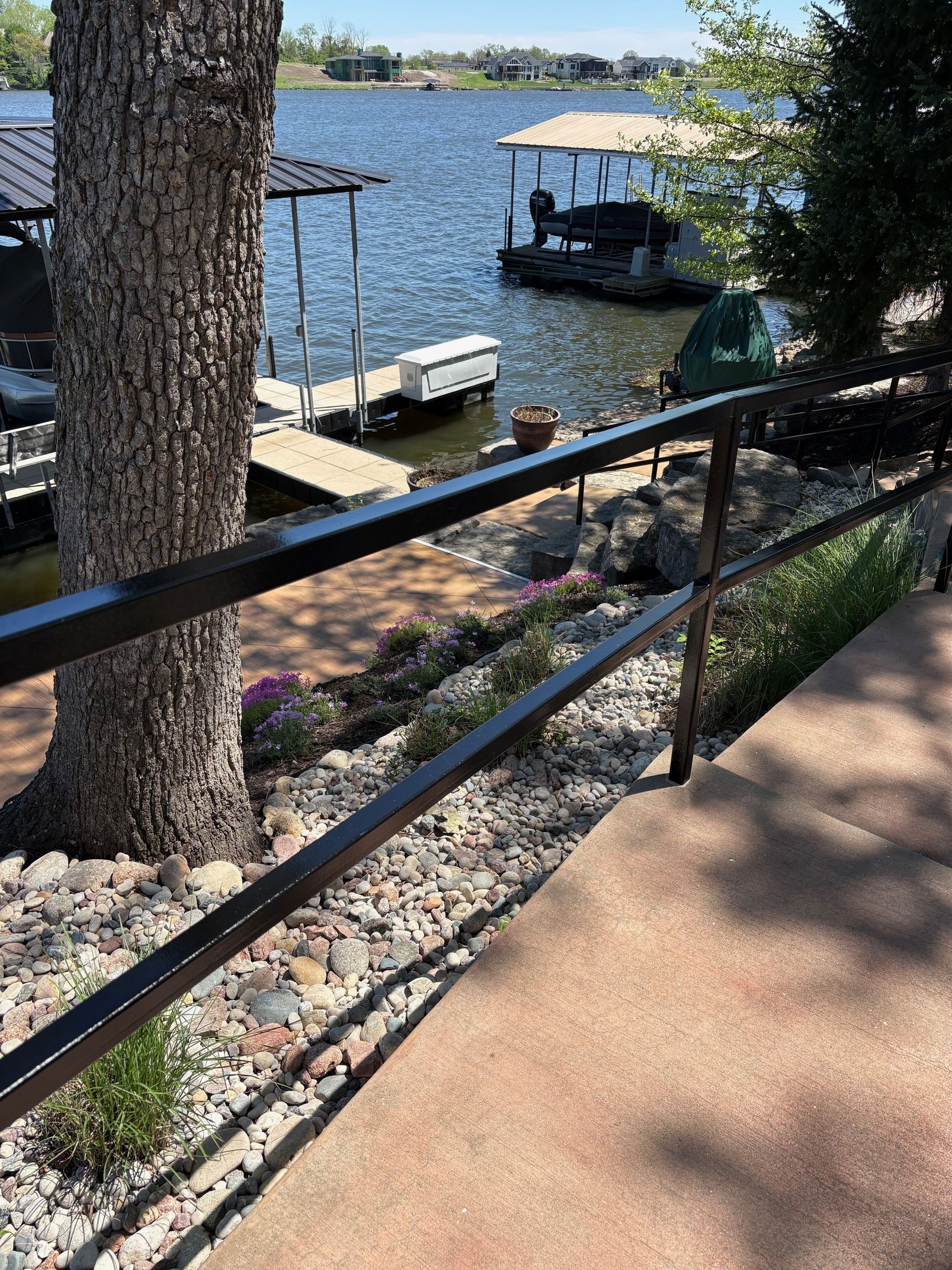 Lakefront dock and boat on a sunny day, viewed past a black railing and rocky shoreline.