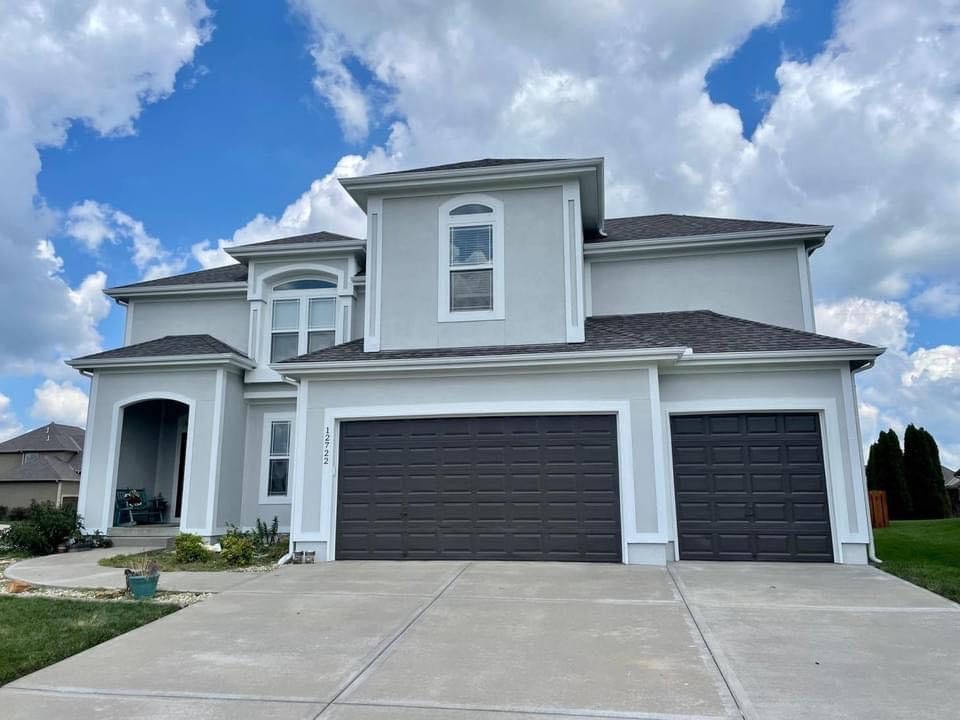 Two-story light gray house with a dark gray three-car garage and arched windows under a blue sky with white clouds.