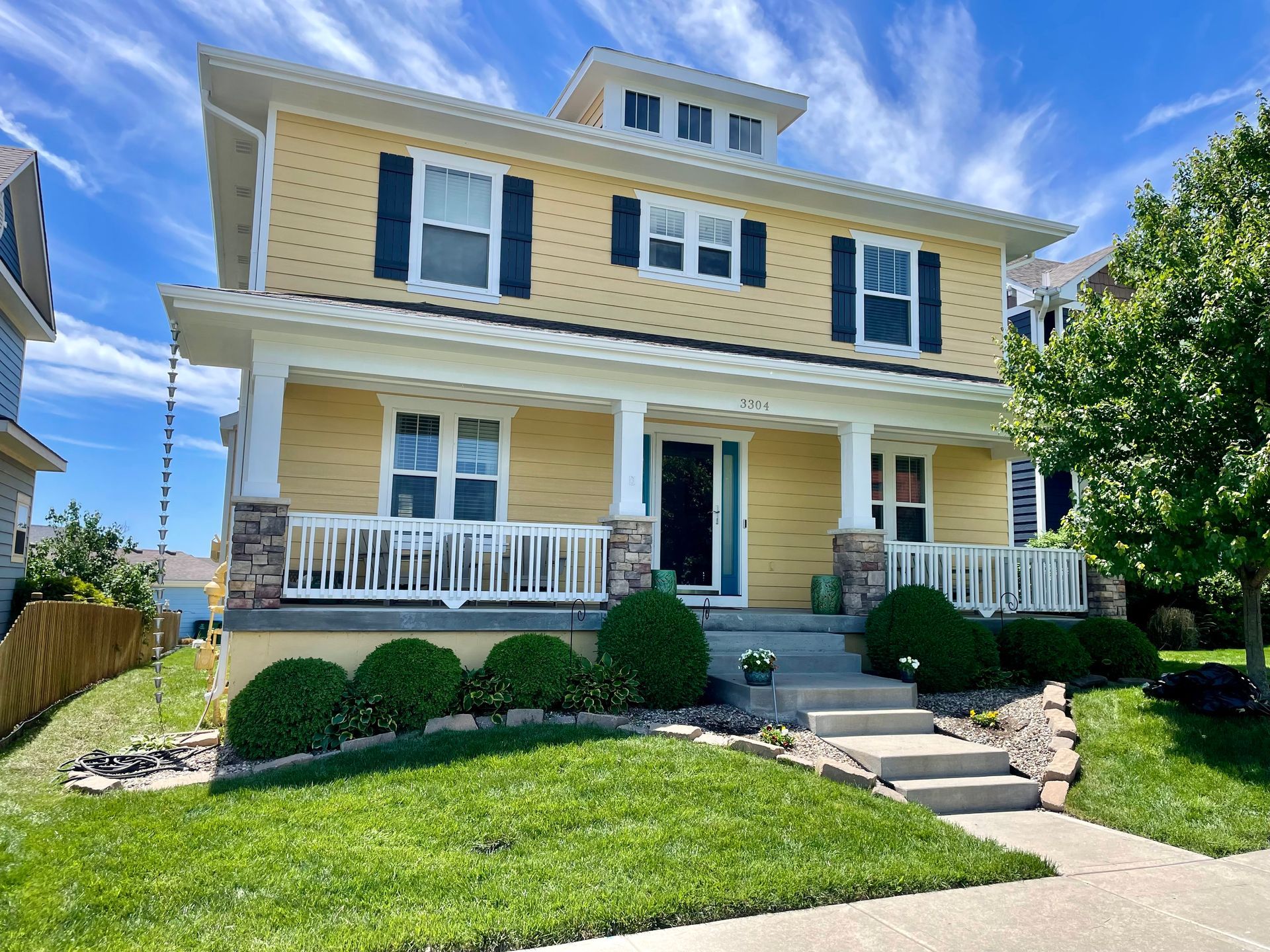 A yellow two-story house with a front porch, white trim, and dark shutters, situated on a grassy lawn under a blue sky.