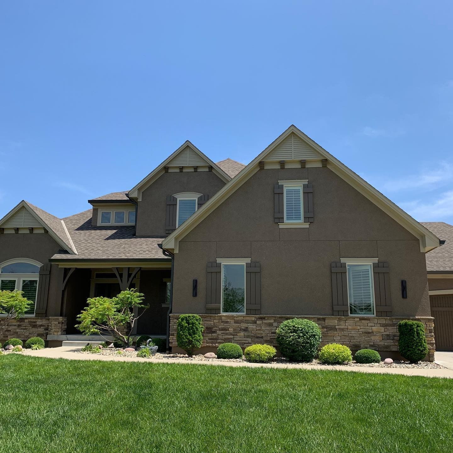 House with brown stucco exterior, stone base, and shutters. Green lawn and blue sky.
