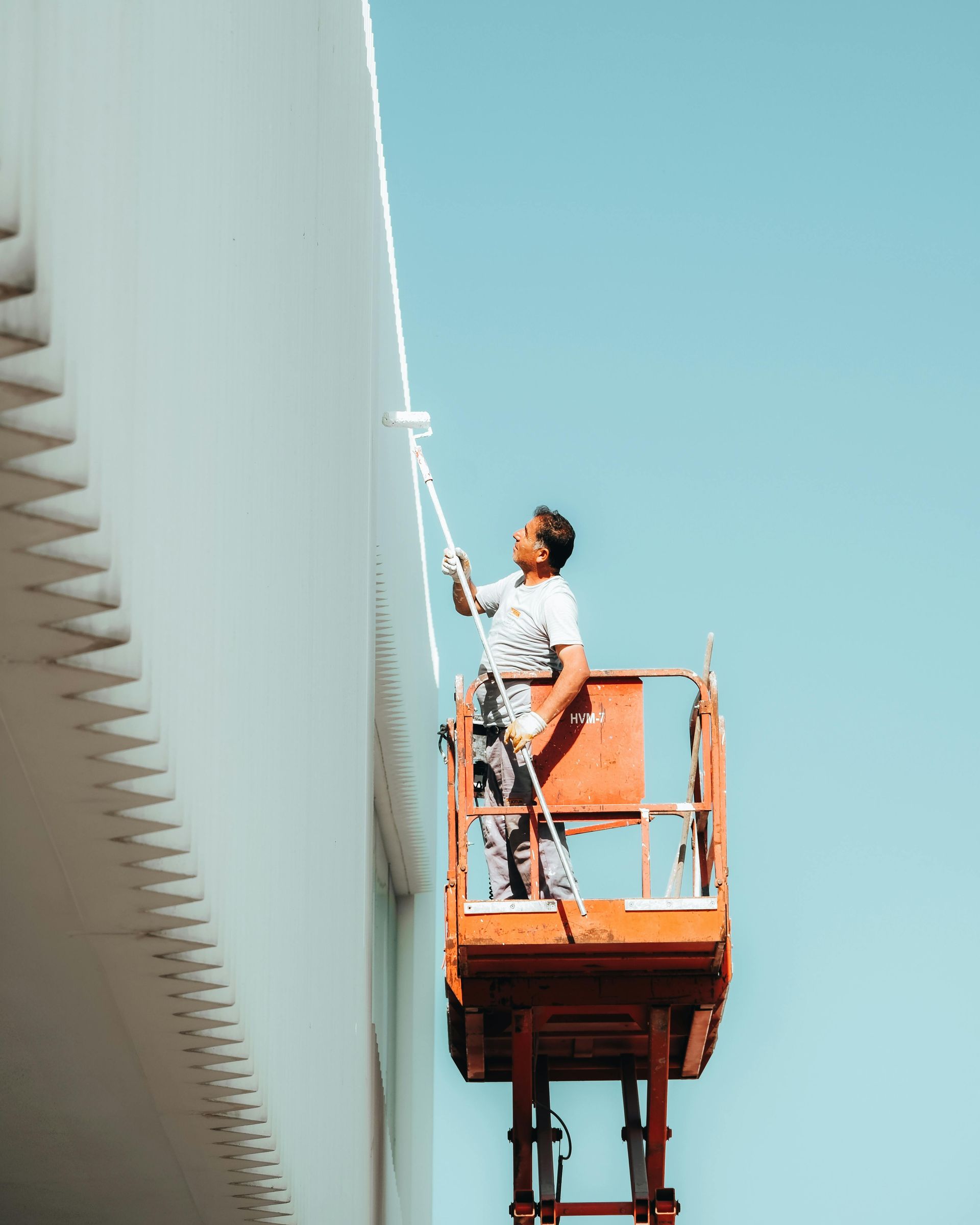 Worker on an orange lift cleaning a tall white building against a blue sky.