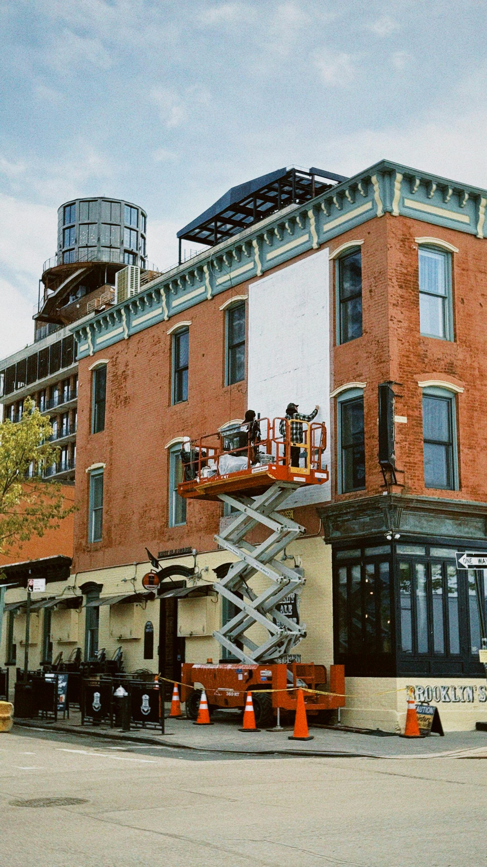 Brick building with workers on a scissor lift repairing the roofline, orange cones below.