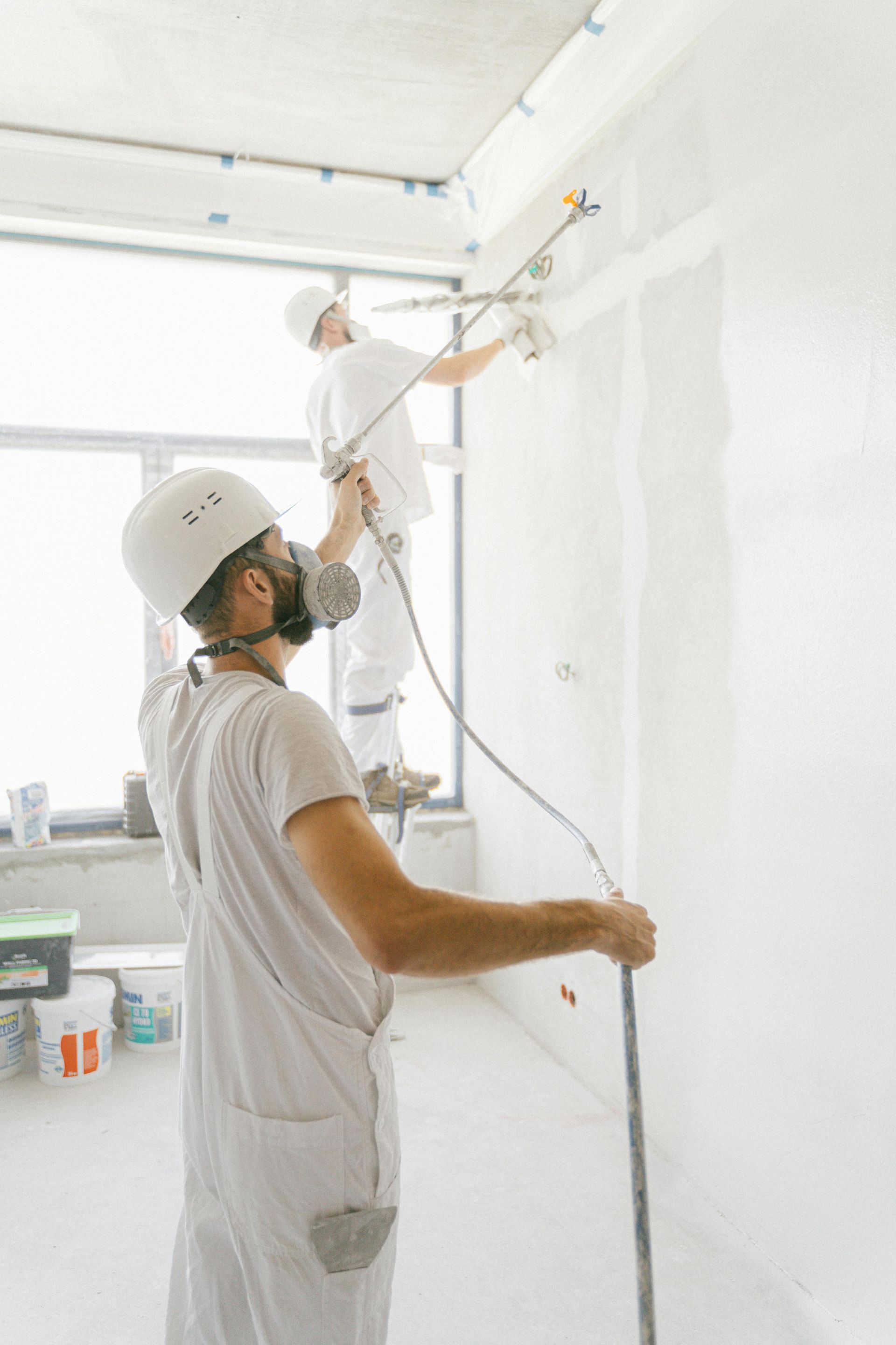 Two painters in white coveralls painting a bright white wall in a sunlit room