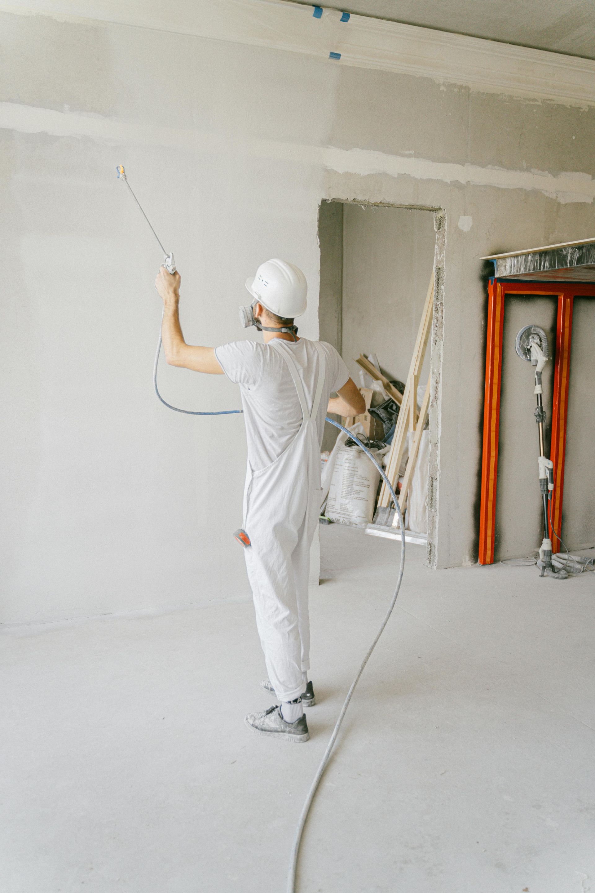 A worker in white protective gear uses a paint sprayer on a wall in a room under construction.