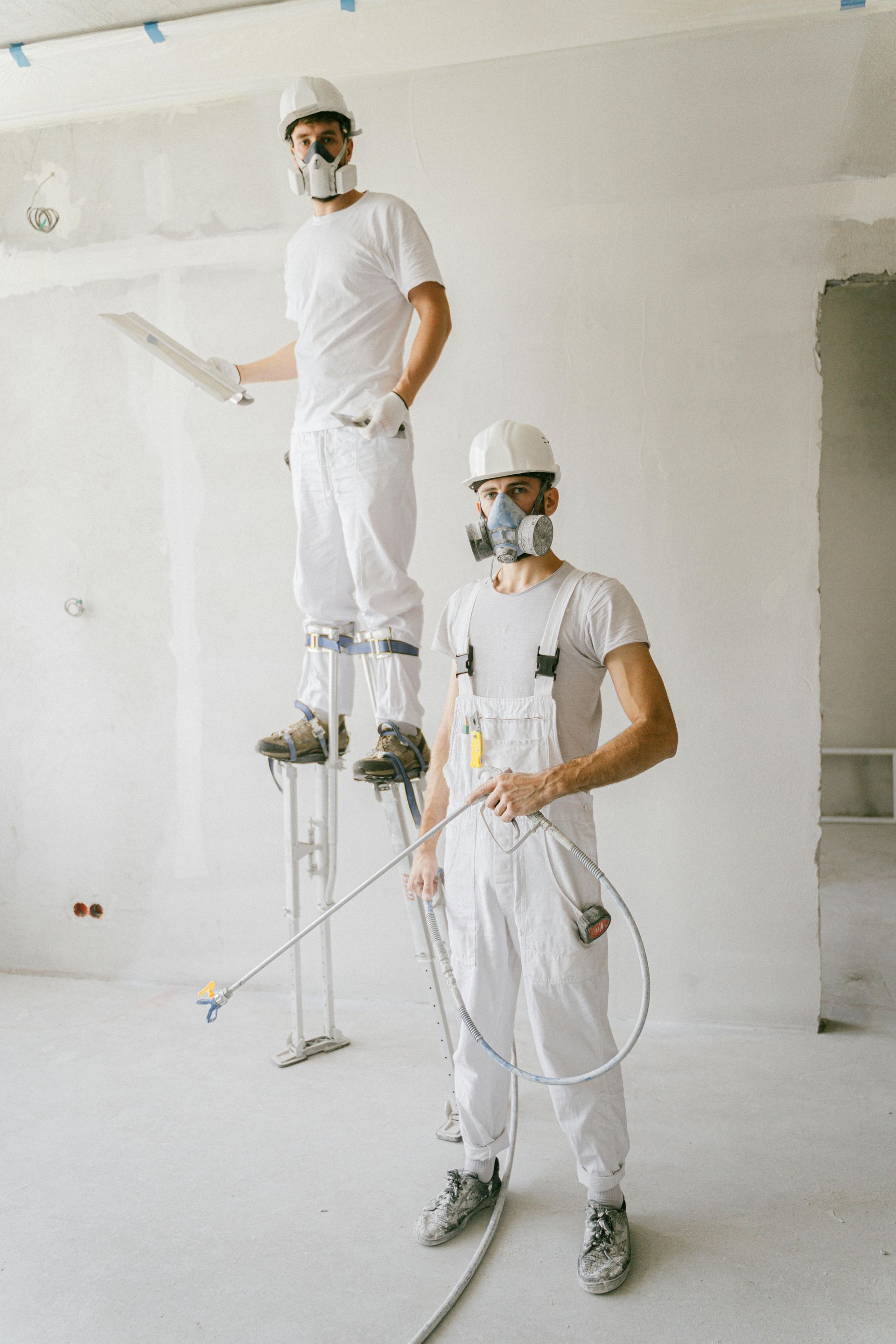 Two painters in white work clothes on metal stilts painting a white wall indoors