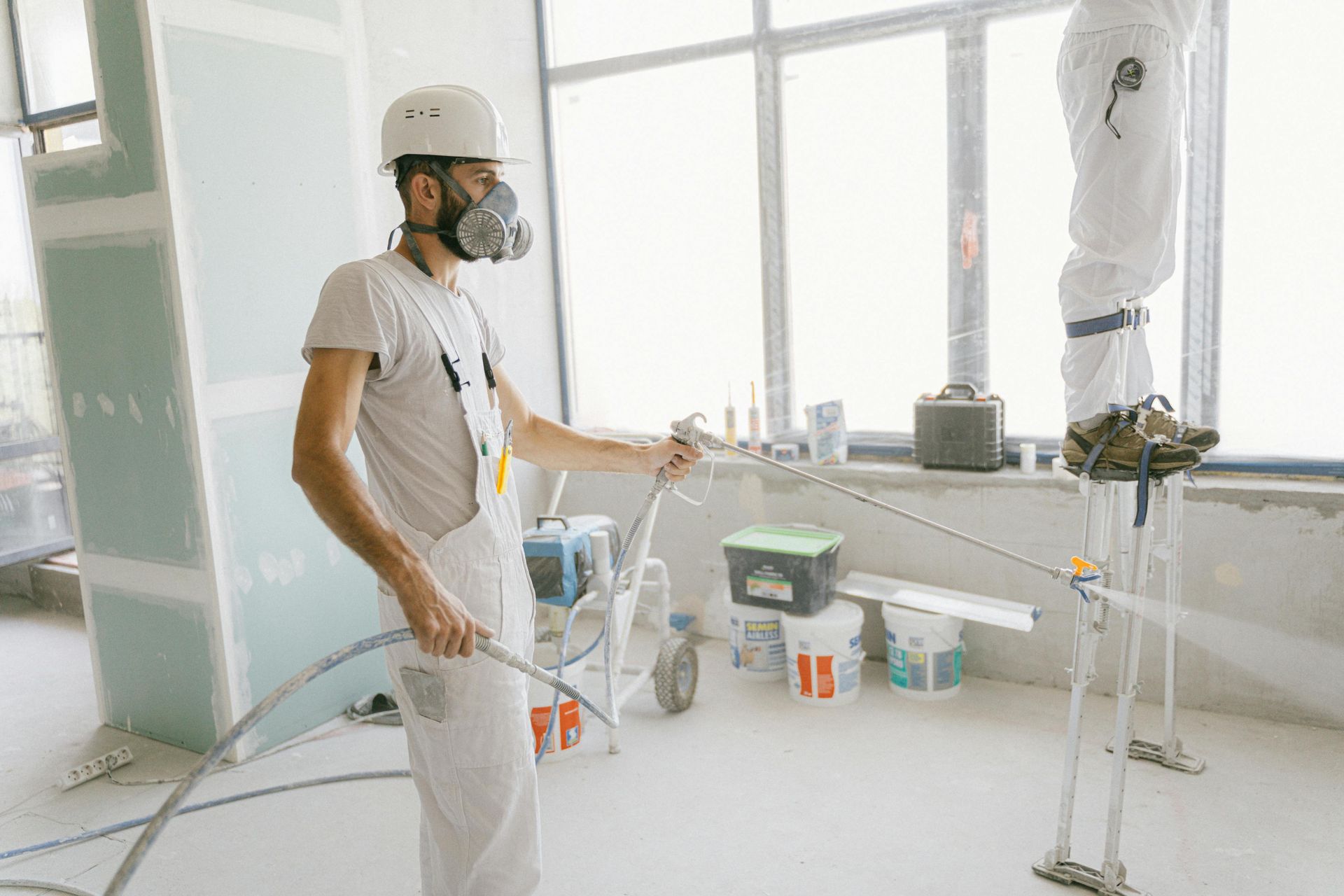 A worker wearing a respirator and white protective gear sprays a wall, while another person stands nearby on stilts.