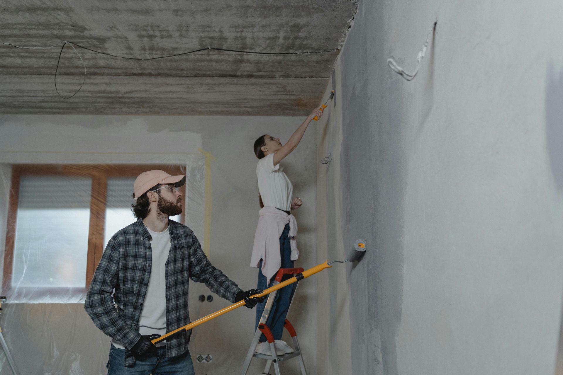A person stands on a ladder while another uses a long-handled roller to paint a gray wall in an unfinished room.