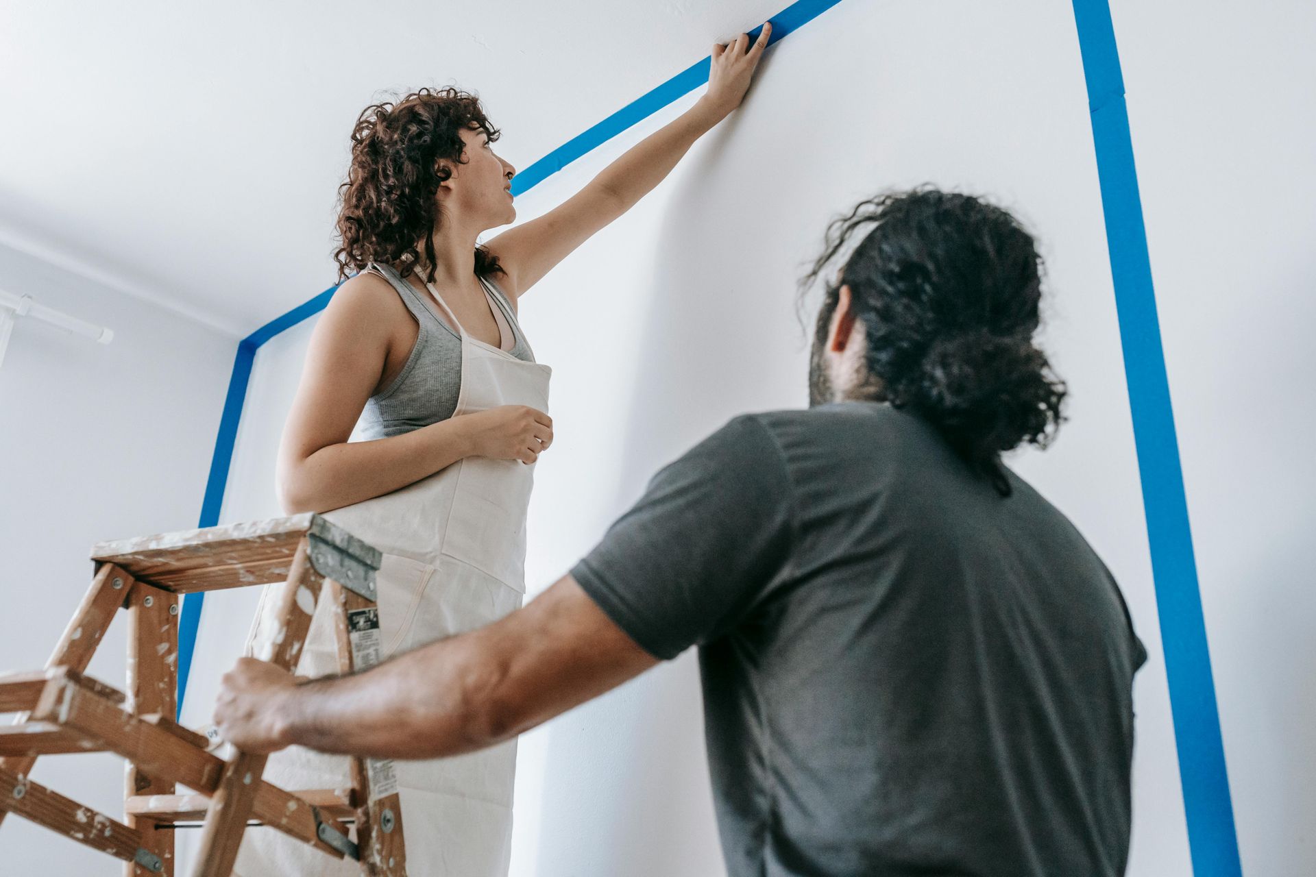 Two people prepare a wall for painting, applying blue painter's tape to form a frame on the white surface.