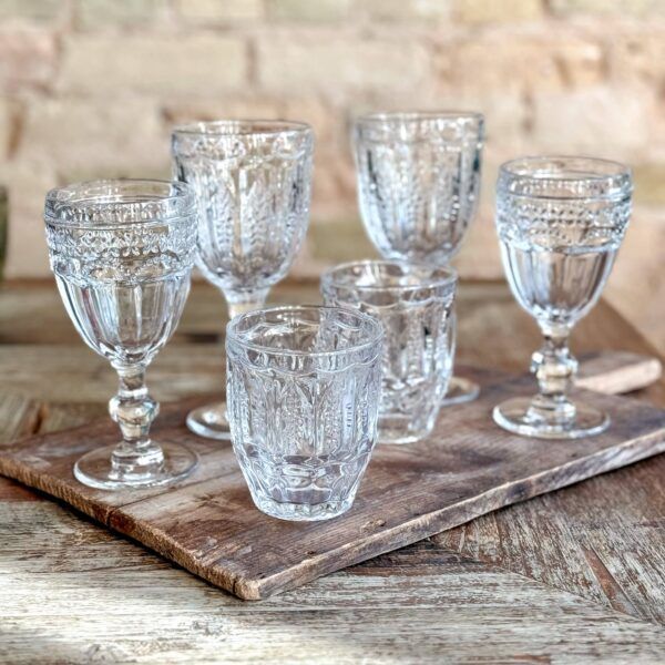 Six clear, ornate glasses on a wooden board, set against a blurred brick wall background.