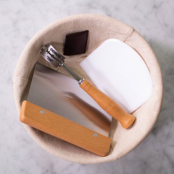 Bread making tools in a round proofing basket: lame, scraper, and dough cutter.