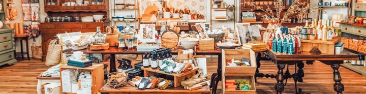 A well-stocked shop with wooden tables displaying various goods. Shelves are filled with items, warm lighting.