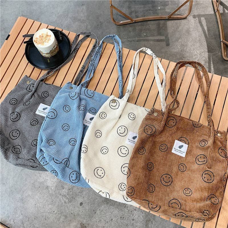 Four corduroy tote bags with smiley faces in gray, blue, cream, and brown on a wooden table.