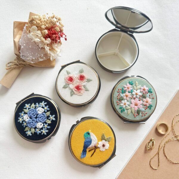 Embroidered compact mirrors in various floral and bird designs, with a small bouquet and jewelry.