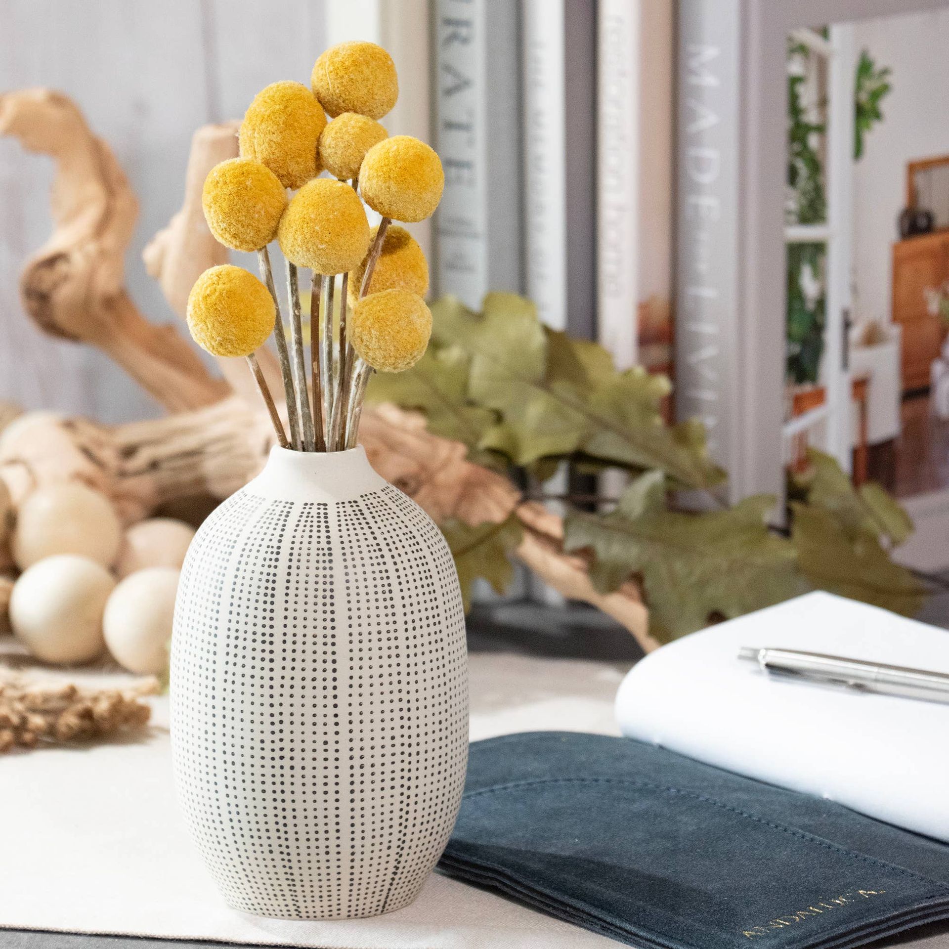 Yellow billy buttons in textured white vase on a desk with books and a notebook.