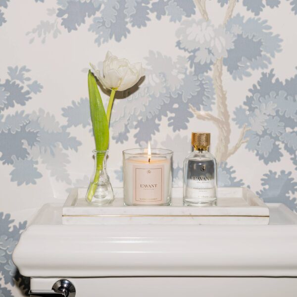 White tulip in vase, lit candle, perfume bottle on tray in front of blue tree wallpaper.
