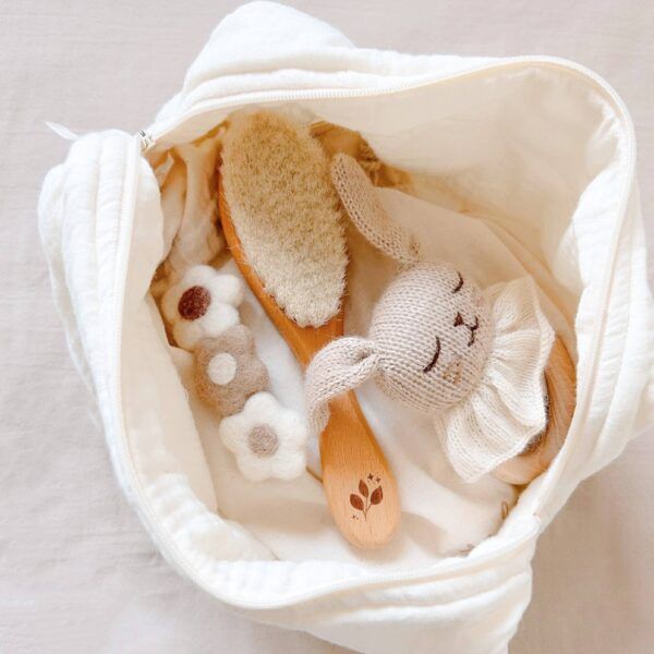 Cream-colored bag open with a wooden brush, knitted bunny rattle, and flower-shaped toys inside.