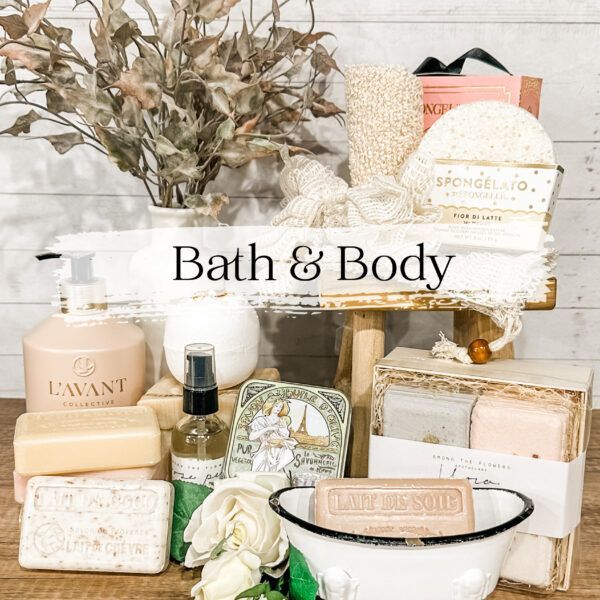Bath and body products arranged on a wooden stool; soaps, loofahs, lotions, spray, and decorative flowers.