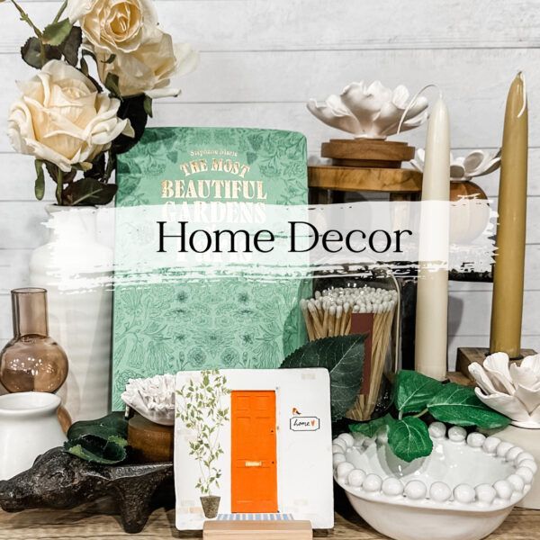 Home decor arrangement with flowers, candles, books, and small orange door on a white wooden background.
