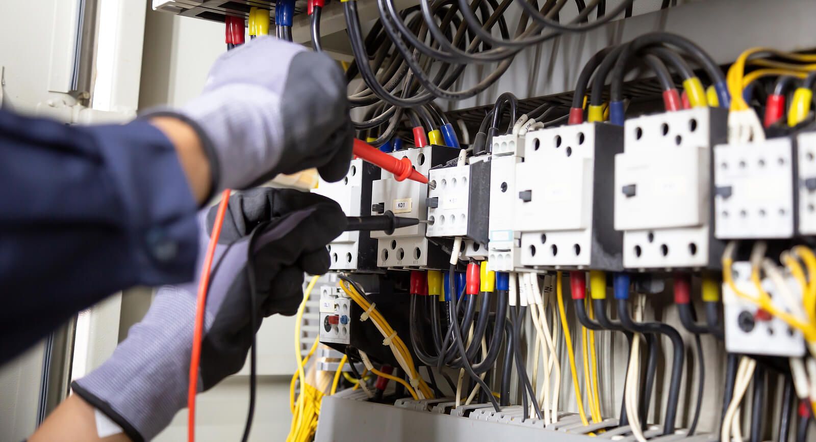 Electrician testing wires in a control panel, wearing gloves, with a multimeter.