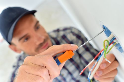 Electrician in a cap, uses a screwdriver to wire a wall outlet; colorful wires visible.