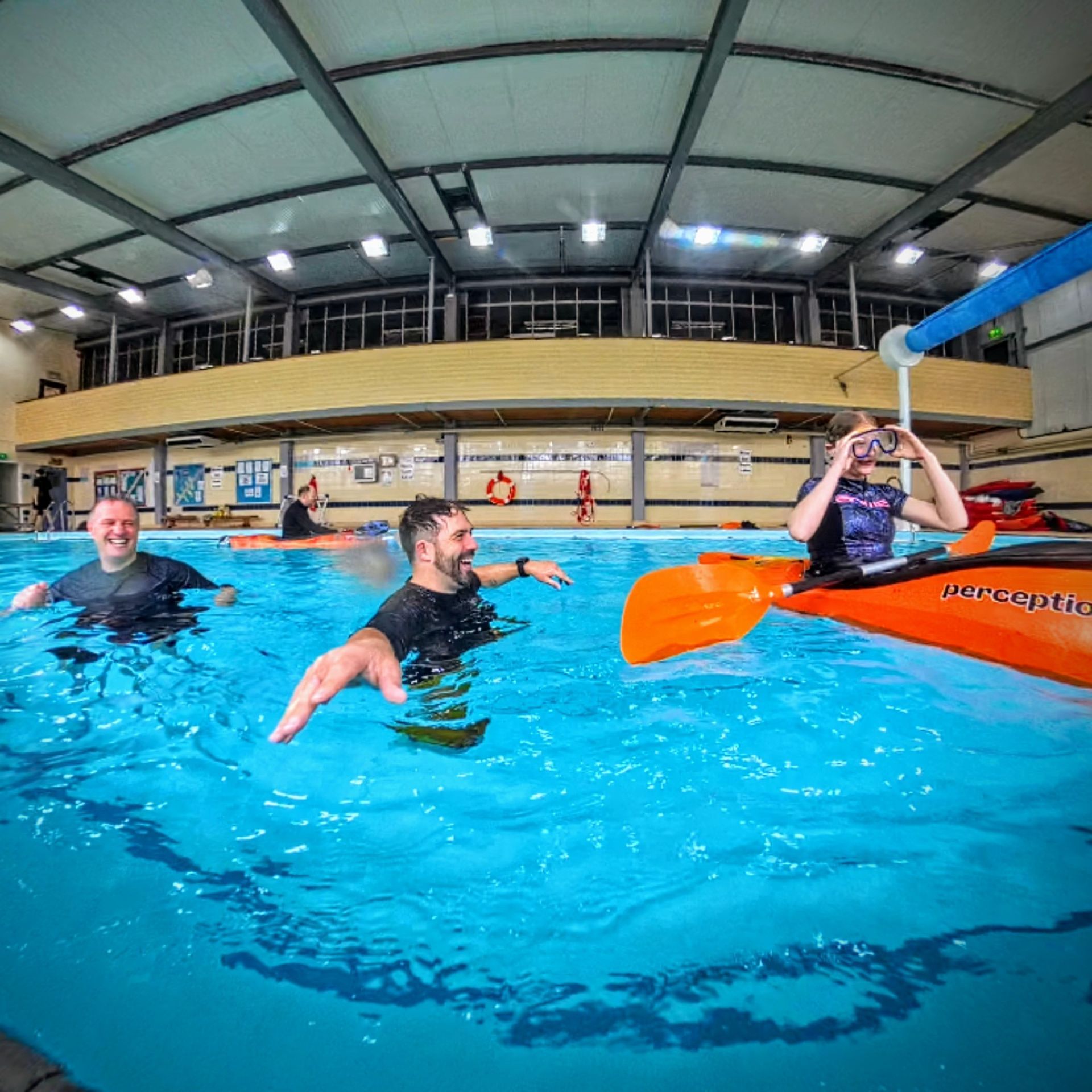 People kayaking in a brightly lit indoor pool. One person is in the kayak, others swim around.
