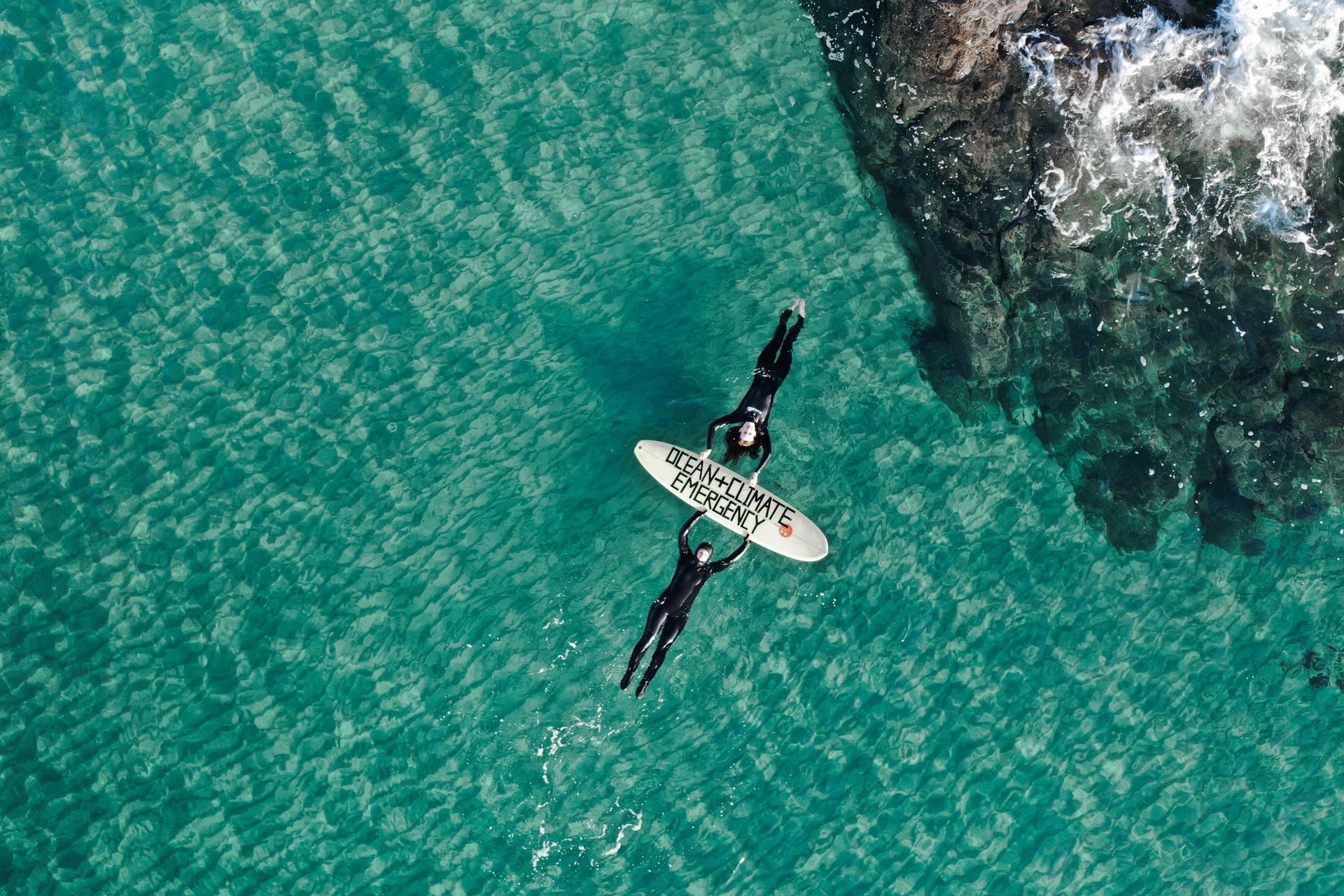 Two people on surfboards in turquoise water near a rocky shore.