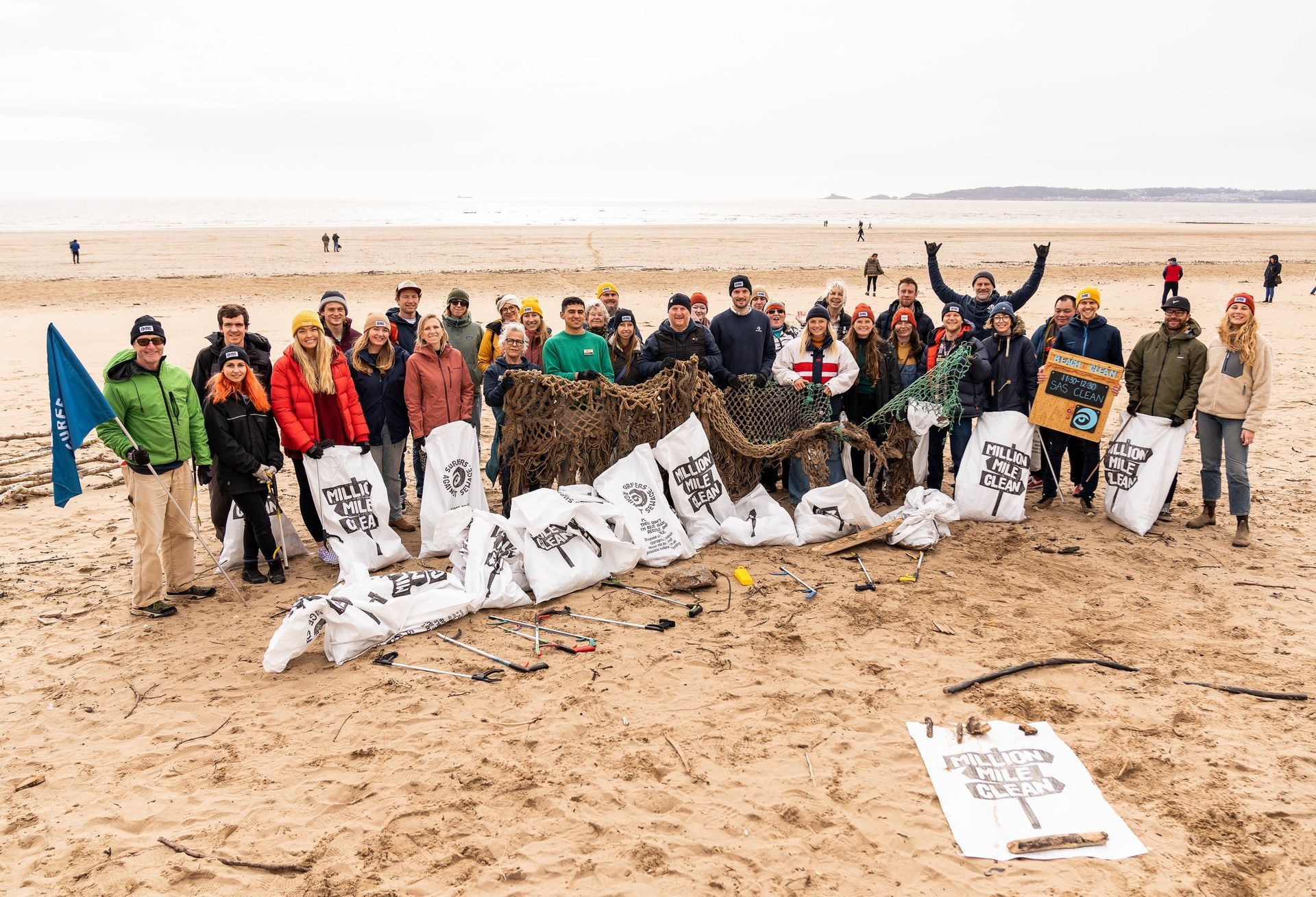 Group of people on a beach holding trash bags after a cleanup. Ocean and overcast sky in background.