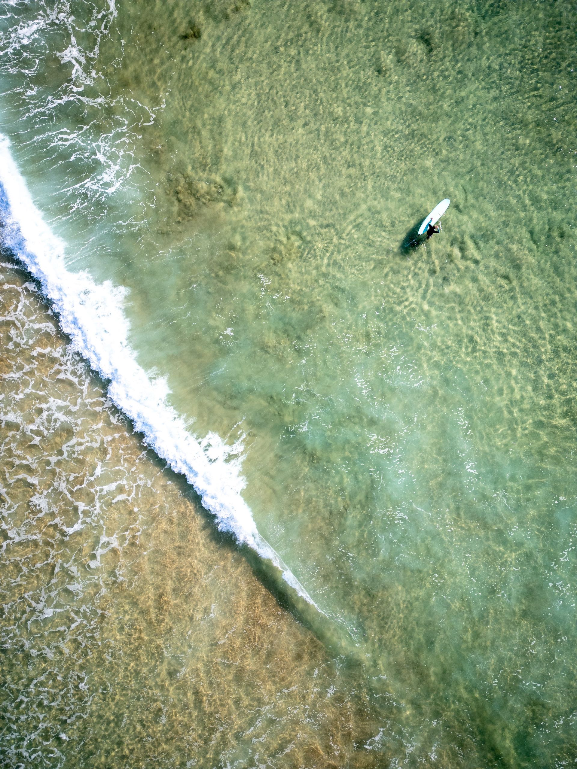 Aerial view: surfer on a longboard in clear turquoise water near the shore with a breaking wave.