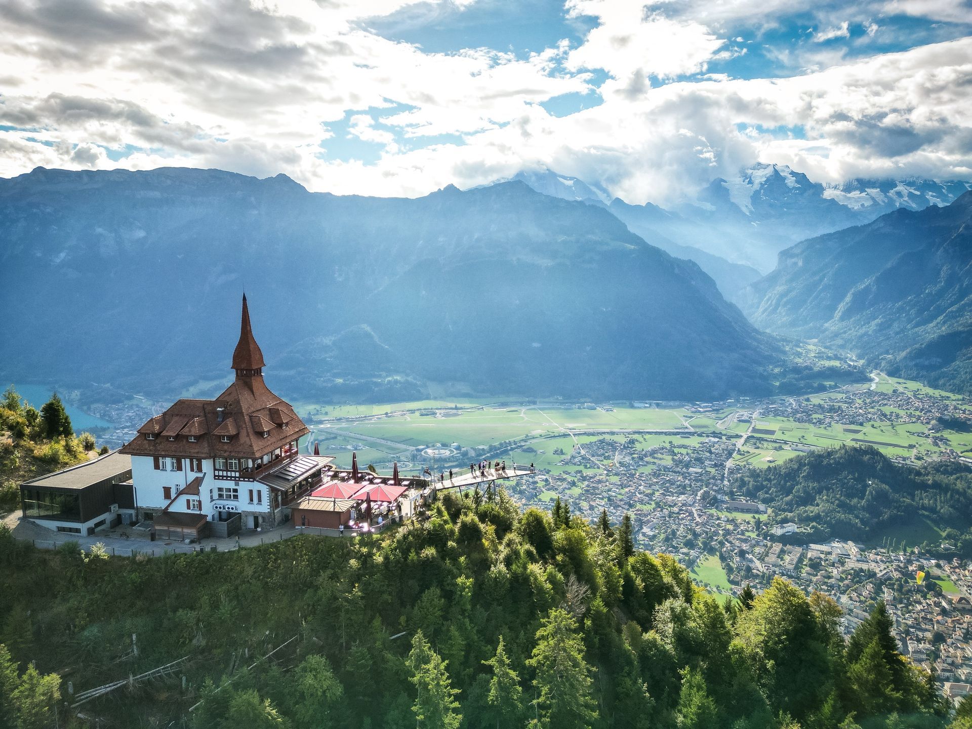 Scenic view of a chalet on a mountain overlooking a valley and distant mountains under a cloudy sky.