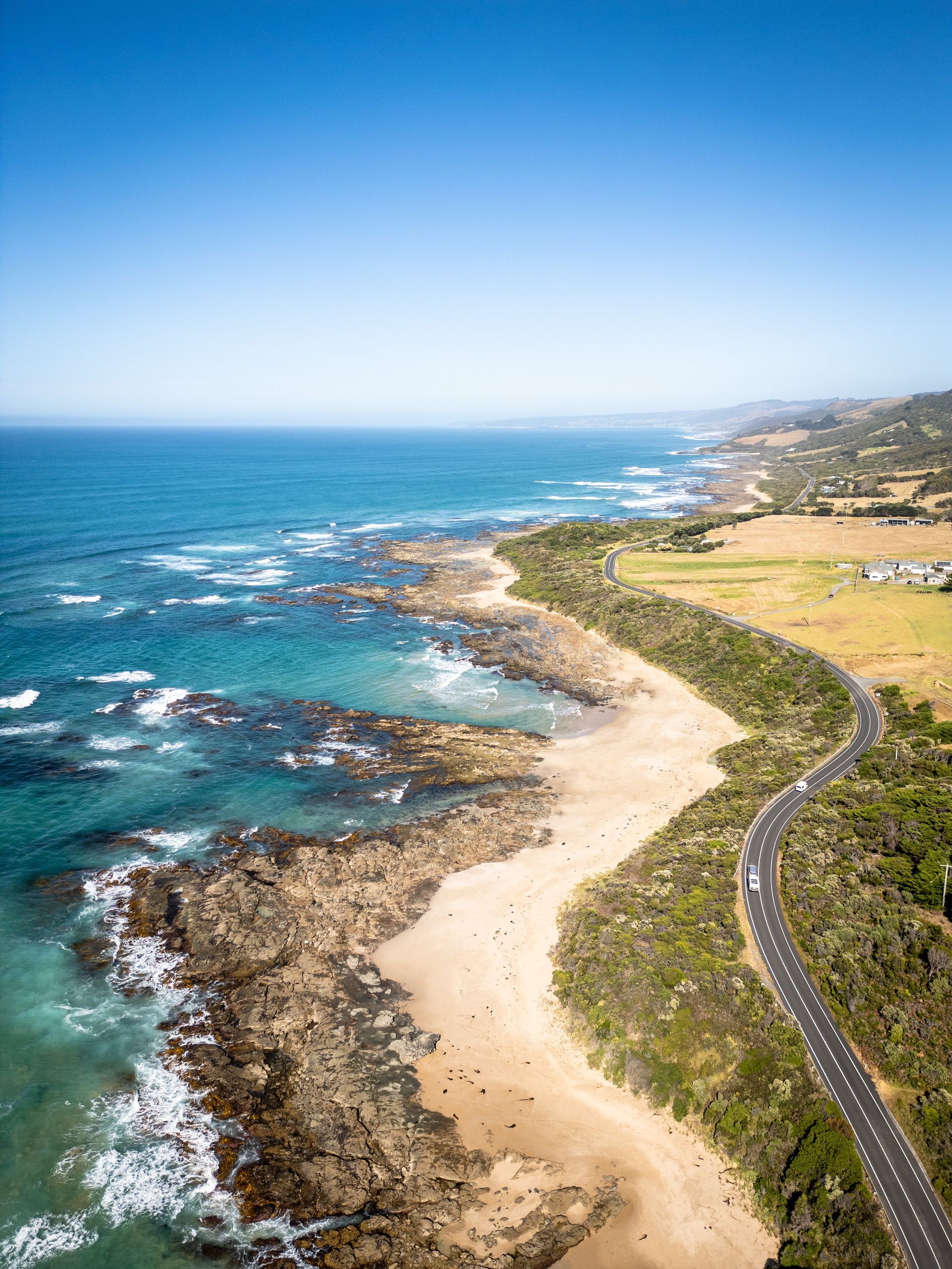 Aerial view of a coastal road winding along a rocky shore, with turquoise water and blue sky.