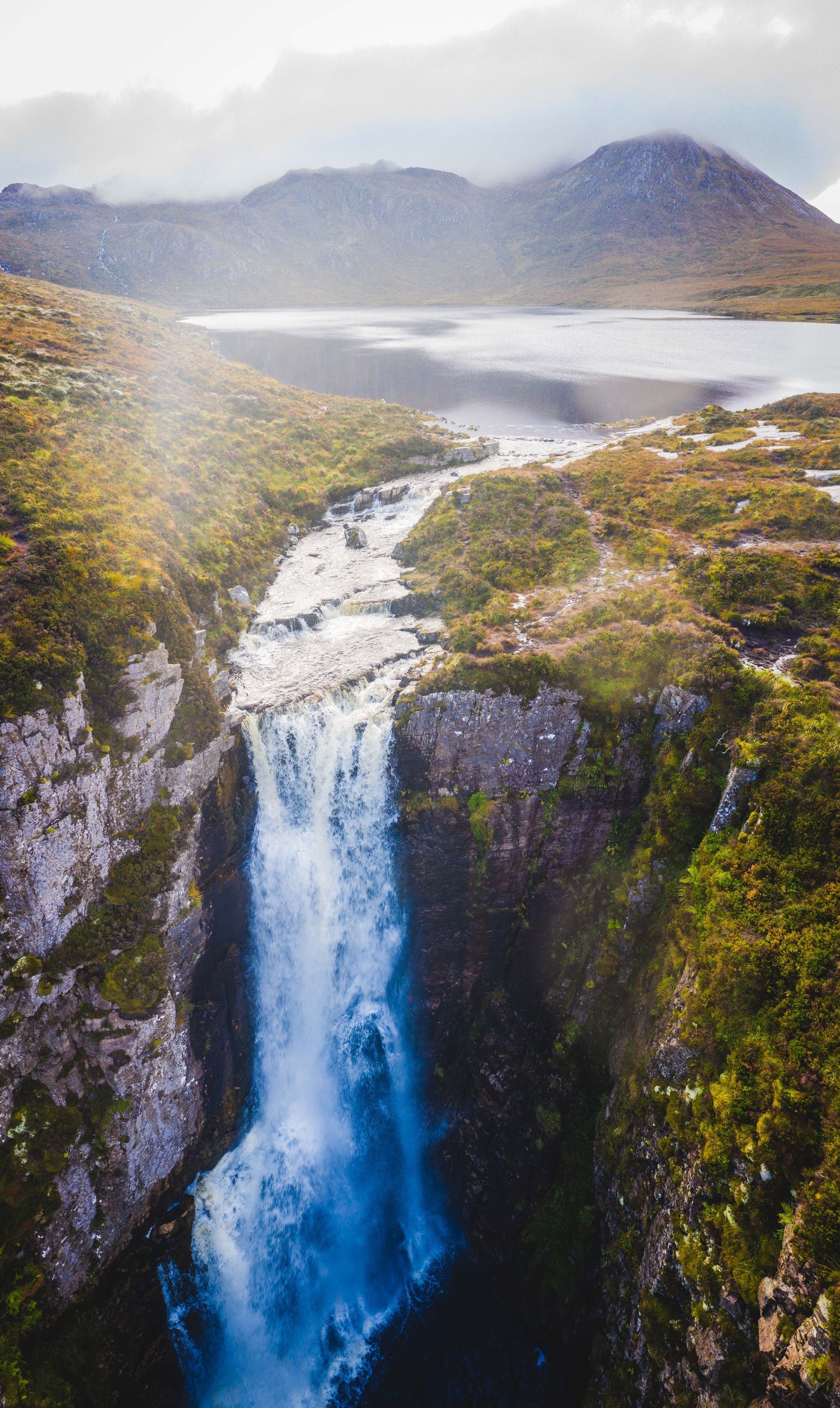 Waterfall cascading down rocky cliffs, leading to a mountain lake. Green and brown vegetation surrounds.