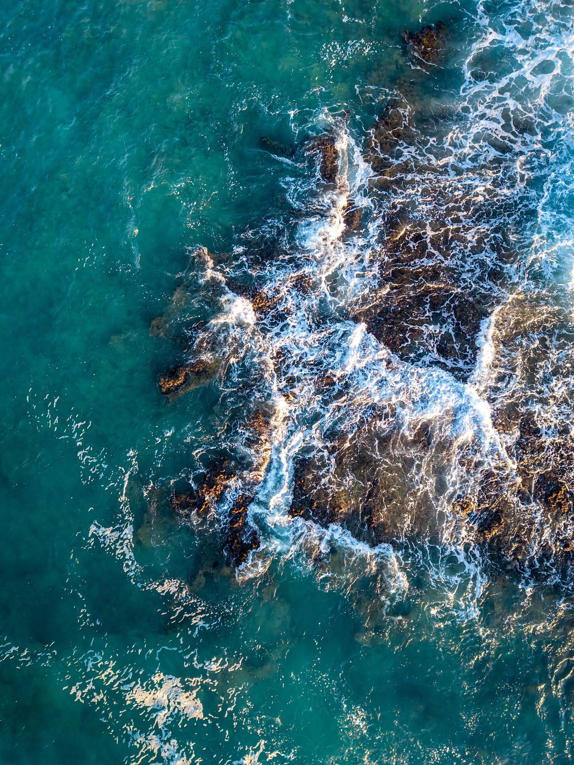 Overhead view of ocean waves crashing against dark rocks in turquoise water.