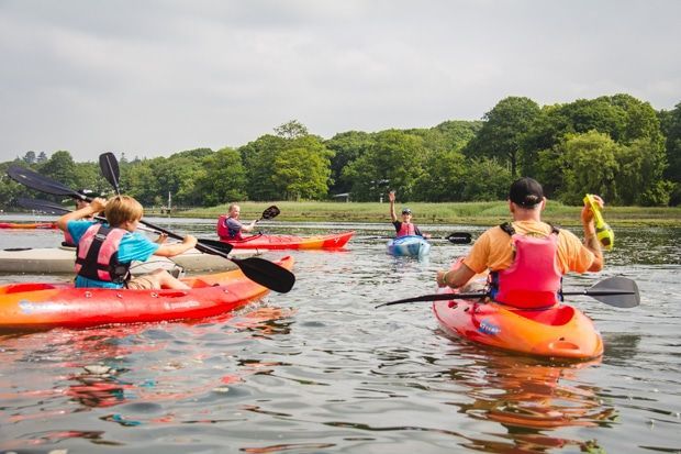 People kayaking on a lake with green trees in the background under a cloudy sky.