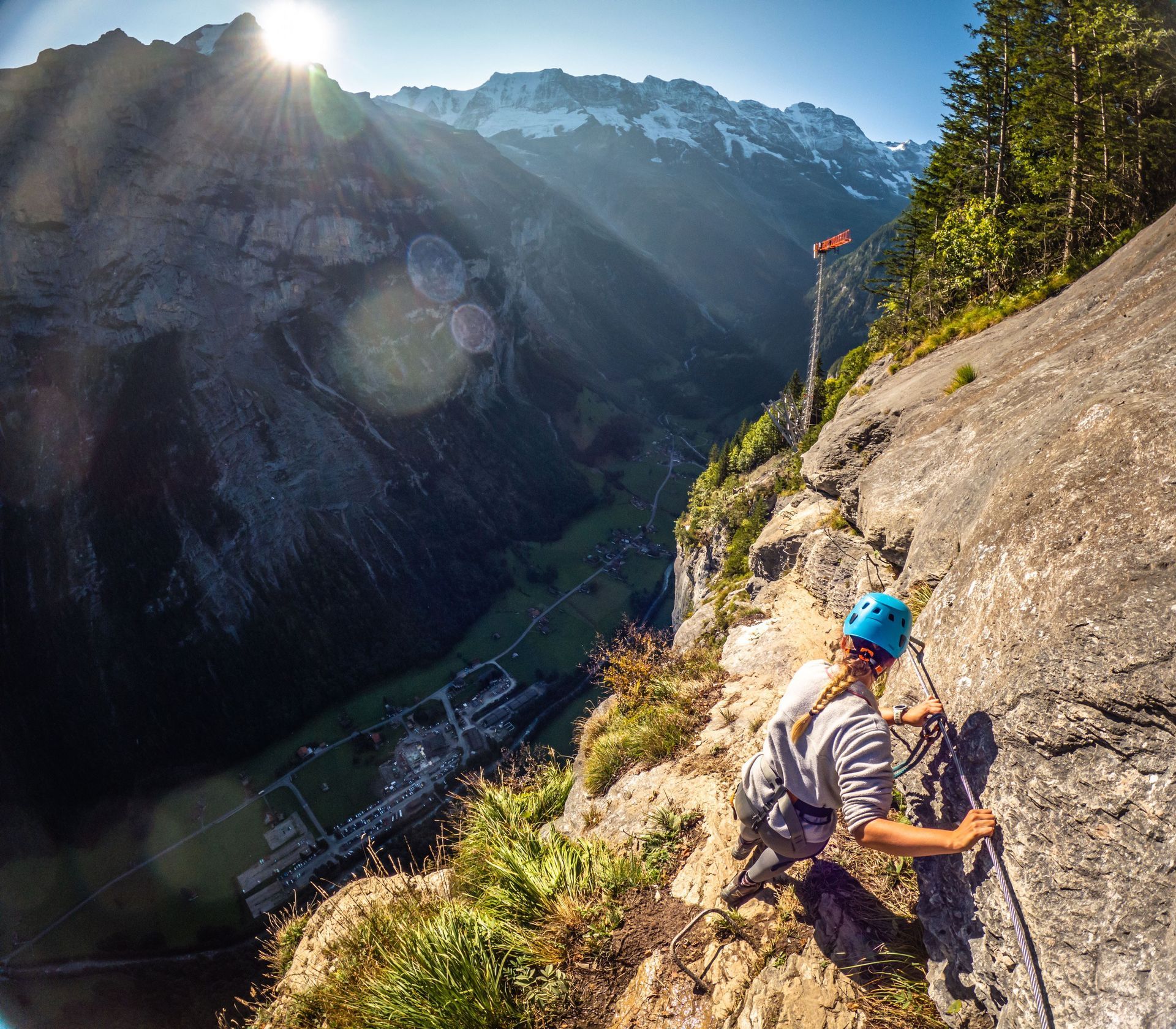 Person climbing a cliffside via ferrata, with a scenic mountain valley in the background.