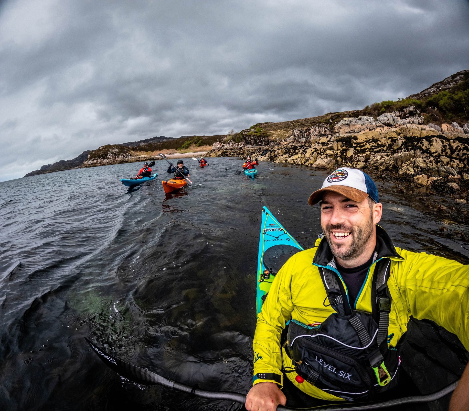 Man smiles while kayaking with a group on a cloudy day. Rocky coastline visible.