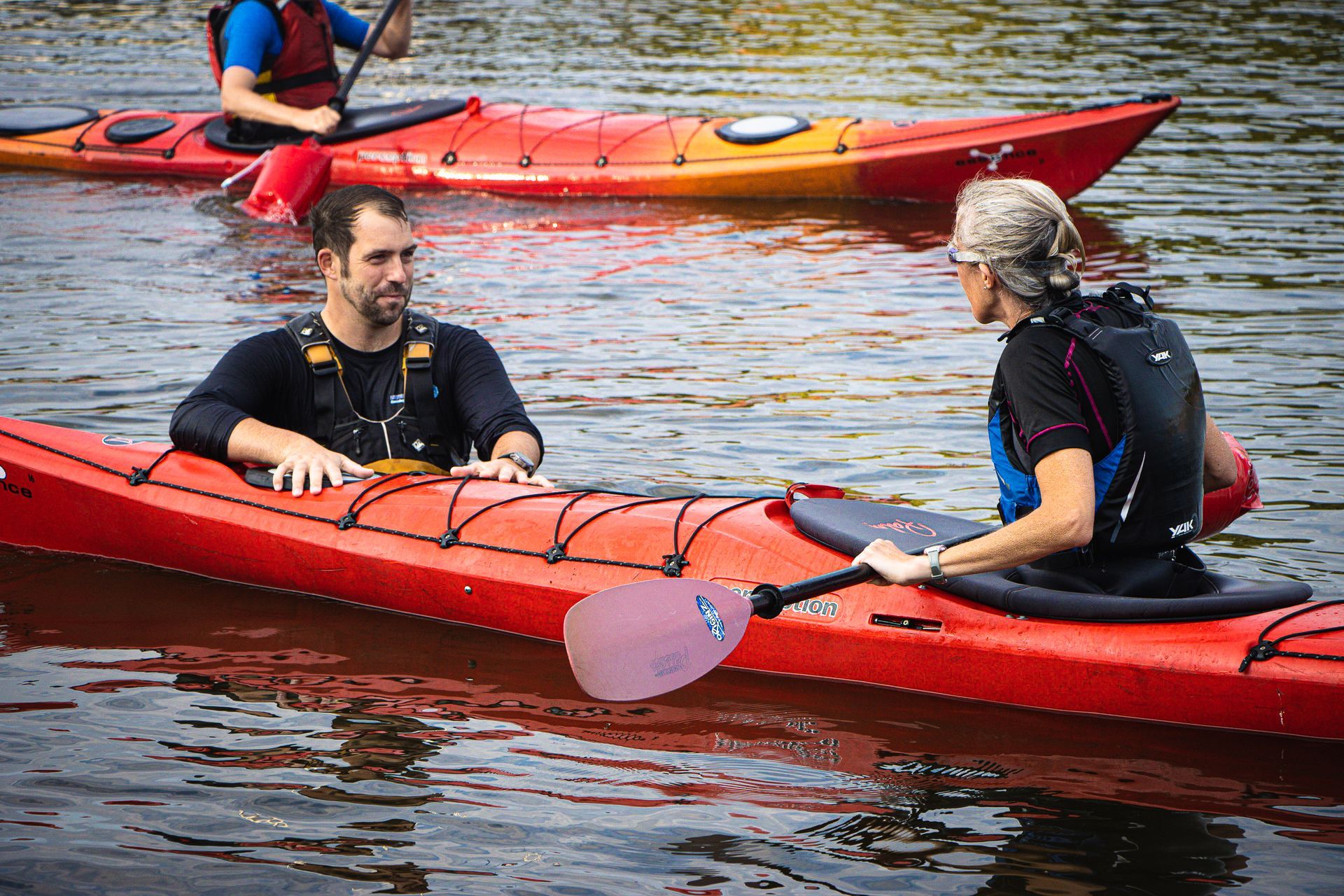 Two people kayaking on water, talking. Red kayaks, one with a paddle.
