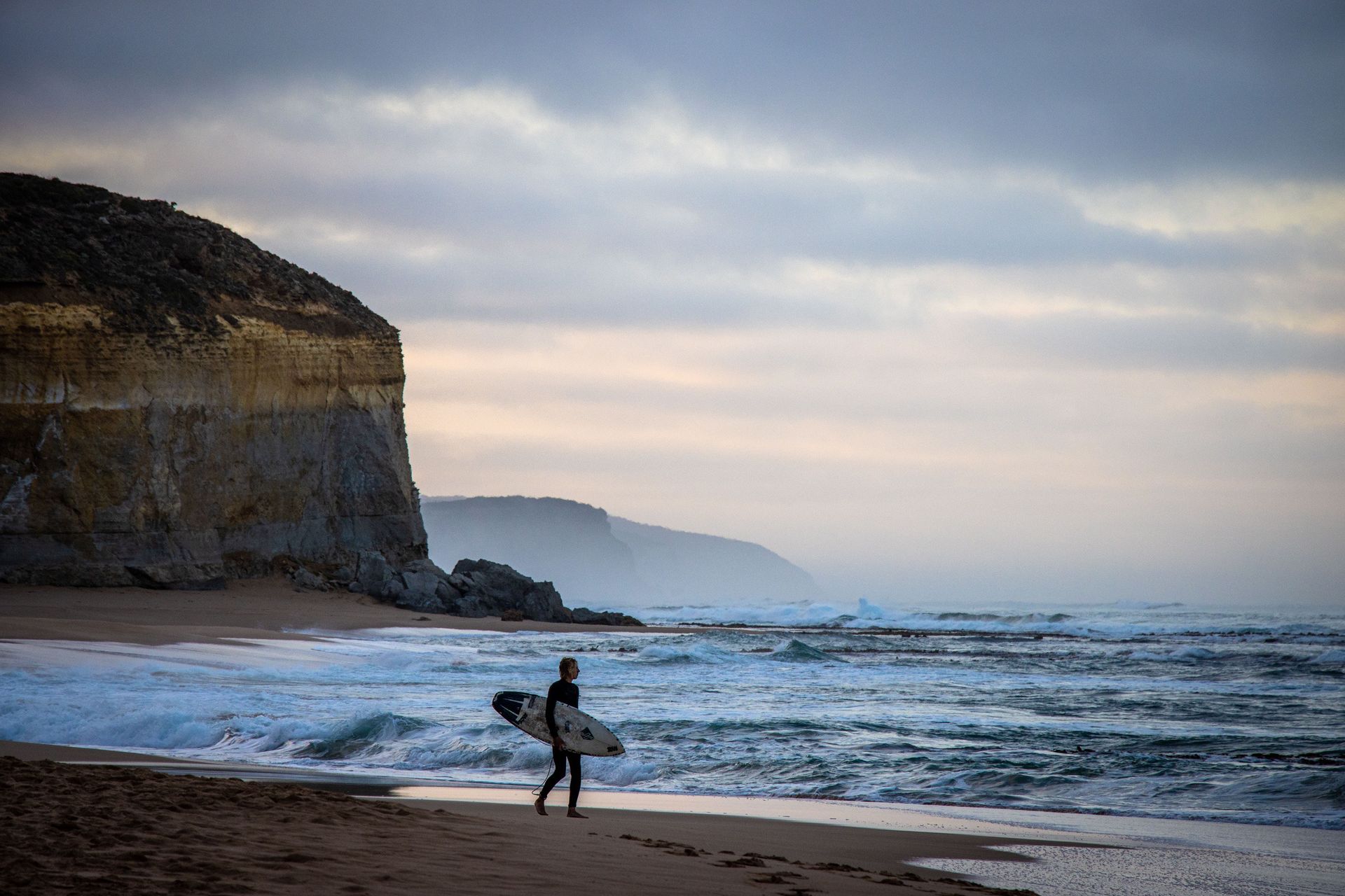 Surfer walks along beach carrying board, cliff backdrop, waves crashing. Overcast sky.